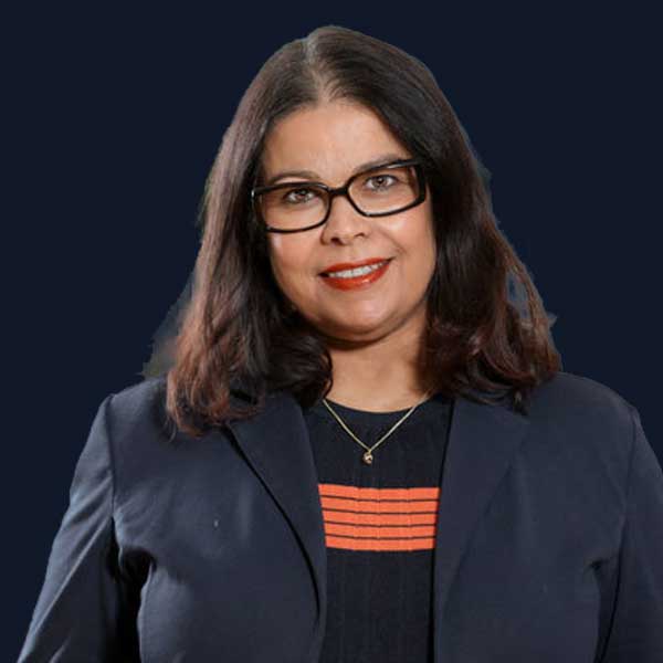 Eine Person mit schulterlangem, dunklem Haar und Brille lächelt und trägt einen marineblauen Blazer über einem dunklen Oberteil mit orangefarbenen Streifen – bereit für ihr nächstes großes Projekt, wie zum Beispiel Ihnen dabei zu helfen, vor einem einfarbigen, dunklen Hintergrund eine „Website erstellen zu lassen“.